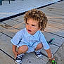 toddler, child, curly_hair, striped_shirt, sneakers, toy, playing, outdoor, dock, wooden_planks, sand, sunlight, casual_clothing, cute, person, young_child, crouching, daylight, fun, curiosity