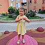 child, girl, yellow_dress, playground, red_platform, outdoor, summer, sandals, greenery, plants, residential_building, windows, sidewalk, people, two_adults, curly_hair, necklace, daylight, smiling, urban