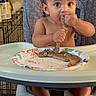 baby, high_chair, sausage, food, plate, eating, child, indoor, curly_hair, messy, wide_eyes, bare_chest, foot, hand, table, household, mealtime, person, seat, expression