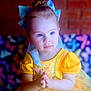 blue_eyes, bow, brick_wall, butterfly_pattern, child, colorful_background, cute, expression, fairy_tale, fashion, girl, hair_bun, hands, indoor, portrait, puffy_sleeves, soft_lighting, toddler, yellow_dress, young