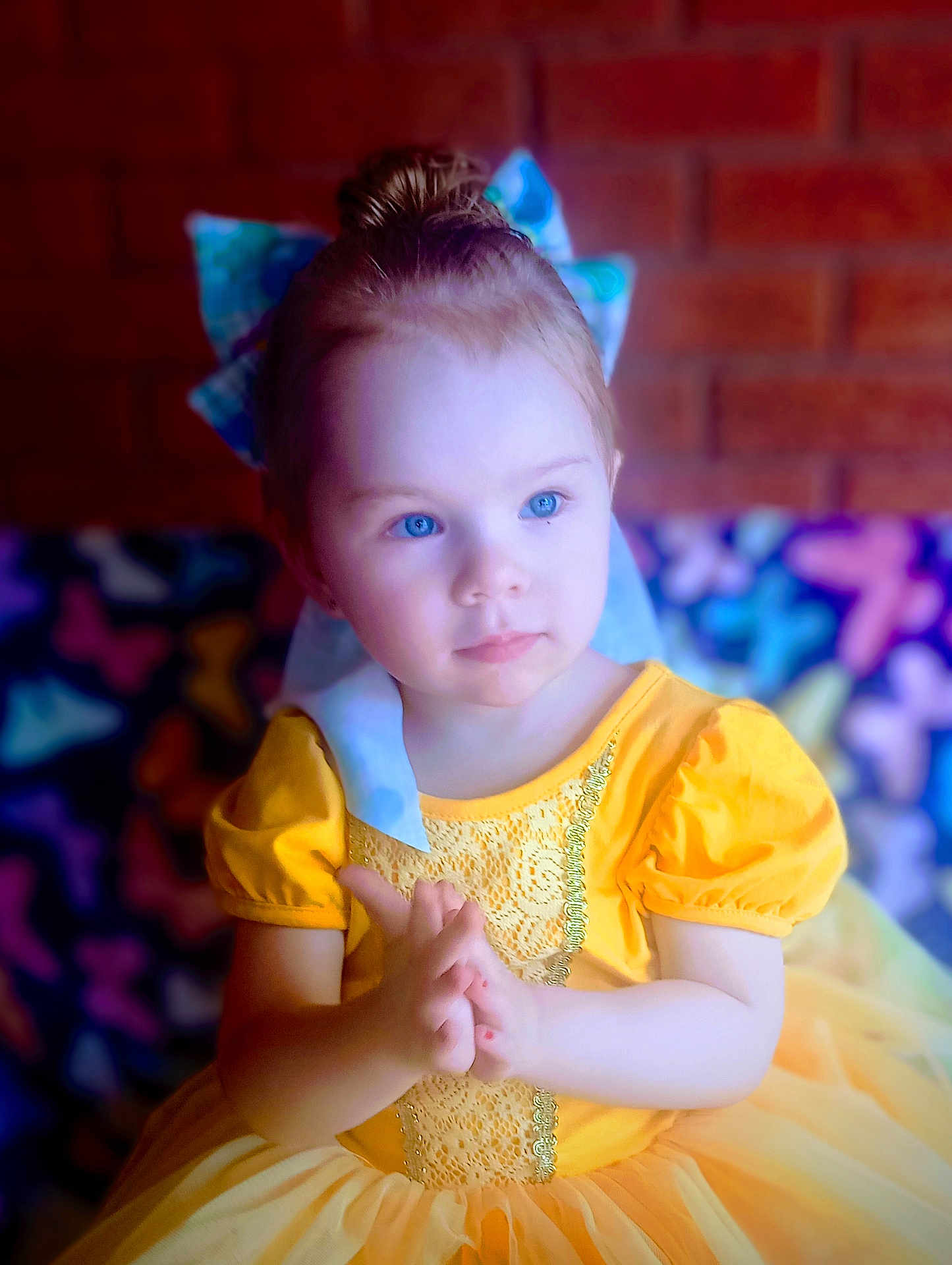 Lue Lue is registered to the contest to win money with this photo: blue_eyes, bow, brick_wall, butterfly_pattern, child, colorful_background, cute, expression, fairy_tale, fashion, girl, hair_bun, hands, indoor, portrait, puffy_sleeves, soft_lighting, toddler, yellow_dress, young