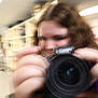 blurred, camera, person, indoor, photography, holding_camera, face, hands, shelves, lighting, room, canon_camera, motion_blur, young_person, technology, focus, equipment, digital_camera, portrait, closeup