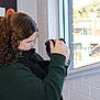 child, curly_hair, camera, indoor, window, person, photography, green_sweater, black_scarf, poster, art, young_person, side_profile, holding_camera, focus, natural_light, building_outside, hair, casual_clothing, brick_wall