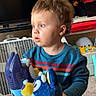 toddler, child, plush_toy, toy, table, indoor, curly_hair, sweater, blue_sweater, stuffed_animal, playroom, carpet, shelf, television, fan, person, cute, curious, young_child, home