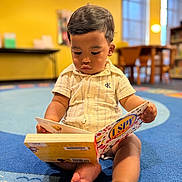 Noah Khalil joined the competition — help win amazing prizes! toddler, child, book, reading, carpet, indoor, yellow_wall, furniture, table, chair, window, soft_lighting, curly_hair, casual_clothing, barefoot, focus, playroom, education, kid, portrait