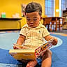 toddler, child, book, reading, carpet, indoor, yellow_wall, furniture, table, chair, window, soft_lighting, curly_hair, casual_clothing, barefoot, focus, playroom, education, kid, portrait