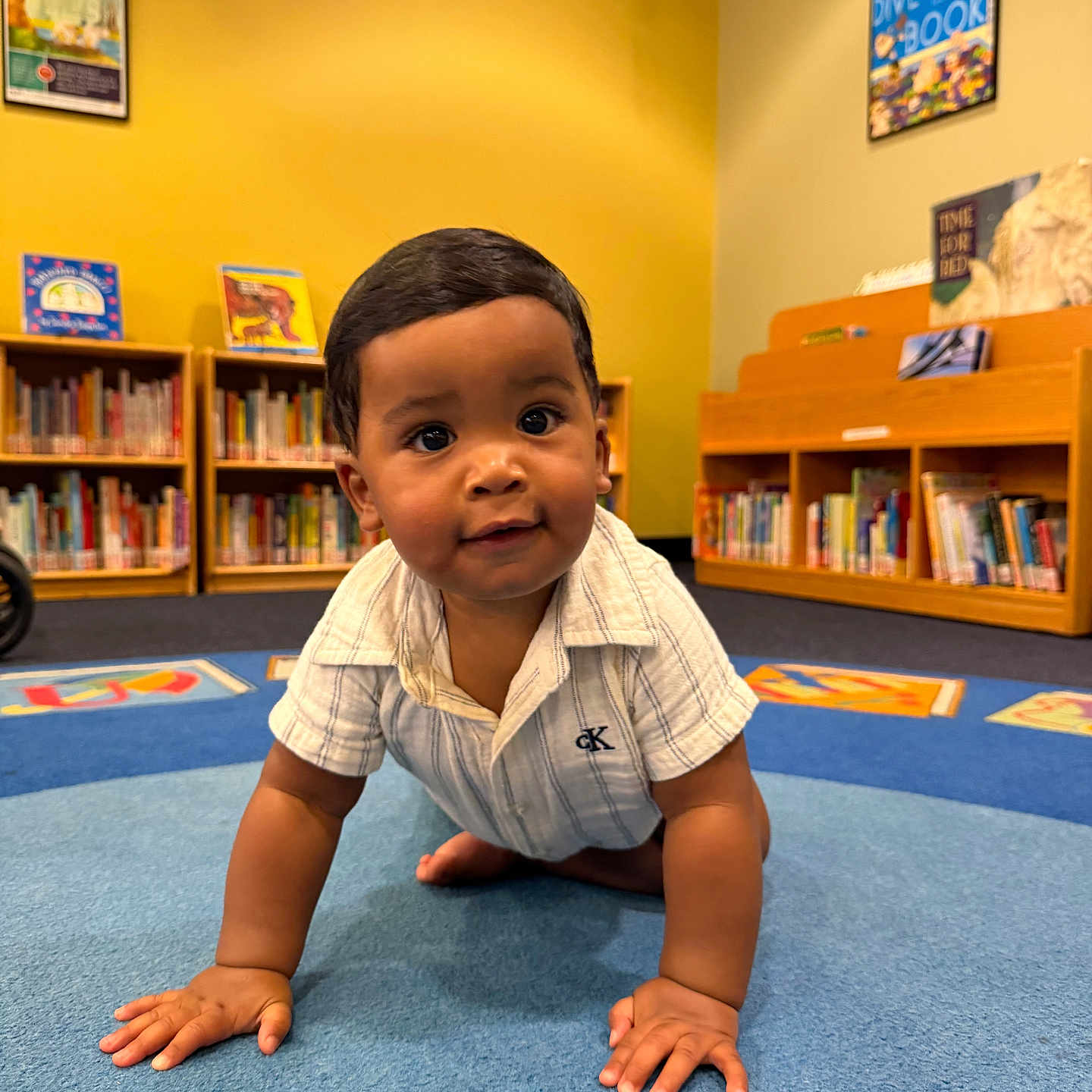Noah Khalil joined the competition — help win amazing prizes! baby, child, crawling, carpet, library, bookshelf, books, yellow_wall, blue_carpet, indoor, curious, smiling, person, infant, short_hair, casual_clothing, floor, young_child, portrait, playful