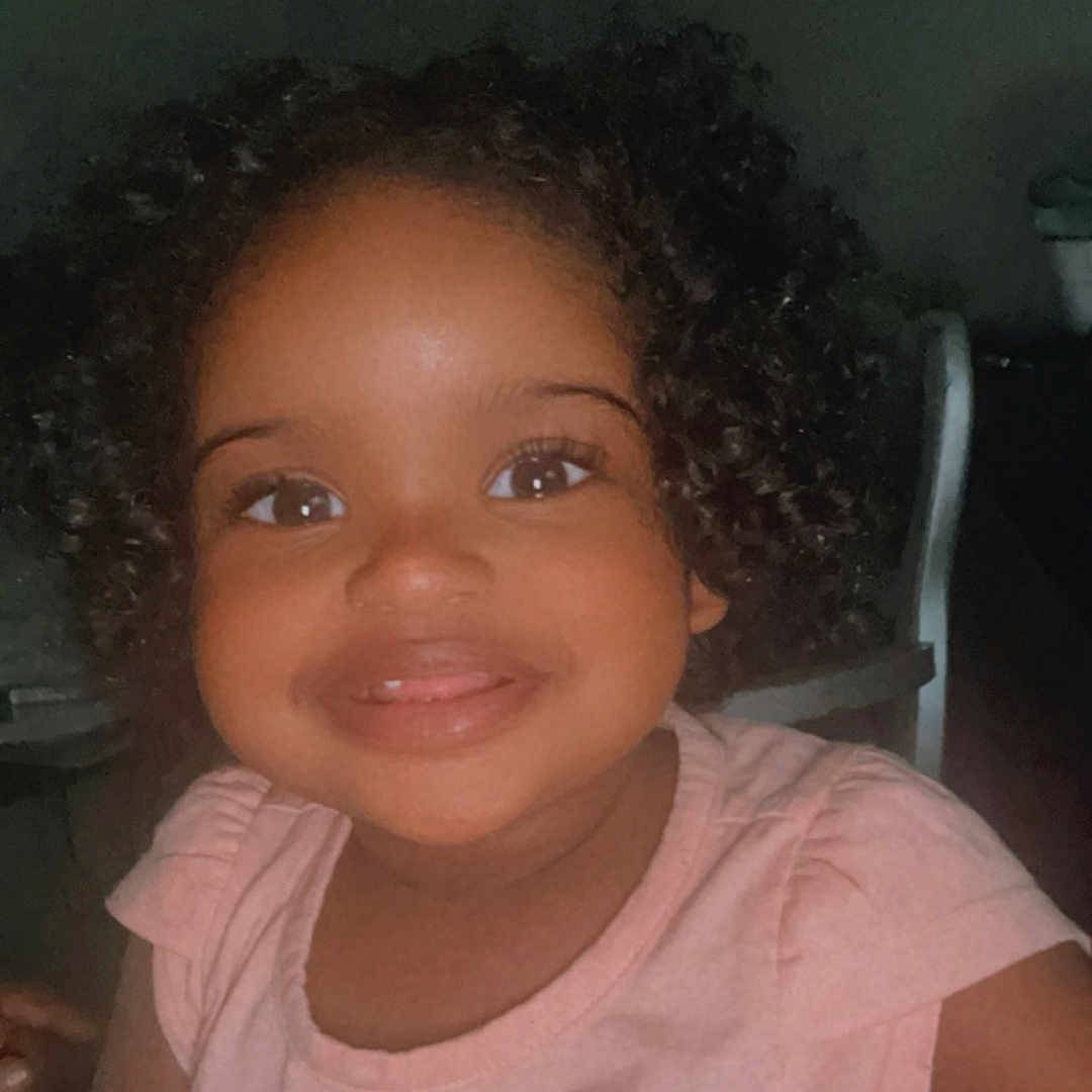 Railyn joined the competition — help win amazing prizes! child, clothing, colorful_text, curly_hair, cute, daisy, dark_background, expression, eyes, face, happy, indoor, light, person, pink_shirt, portrait, sitting, smiling, toddler, young_child