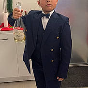 Tony-Junior a rejoint le concours — aidez-le/la à gagner de superbes lots ! boy, child, suit, bow_tie, champagne_bottle, kitchen, holiday_decor, serious_expression, standing, floor, cabinet, door, festive, young, indoors, person, footwear, celebration, portrait, holding