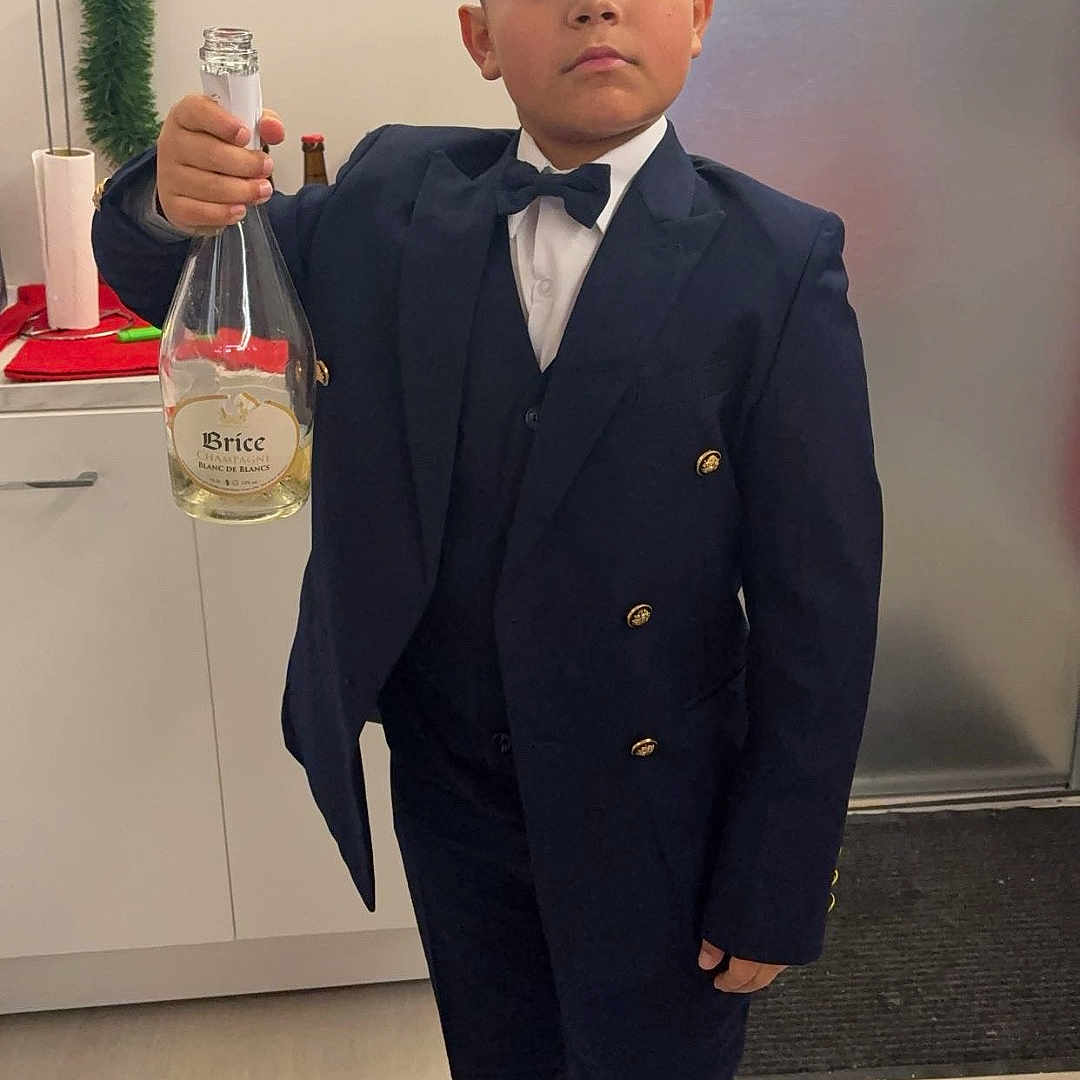 Tony-Junior a rejoint le concours — aidez-le/la à gagner de superbes lots ! bow_tie, boy, cabinet, celebration, champagne_bottle, child, door, festive, floor, footwear, holding, holiday_decor, indoors, kitchen, person, portrait, serious_expression, standing, suit, young