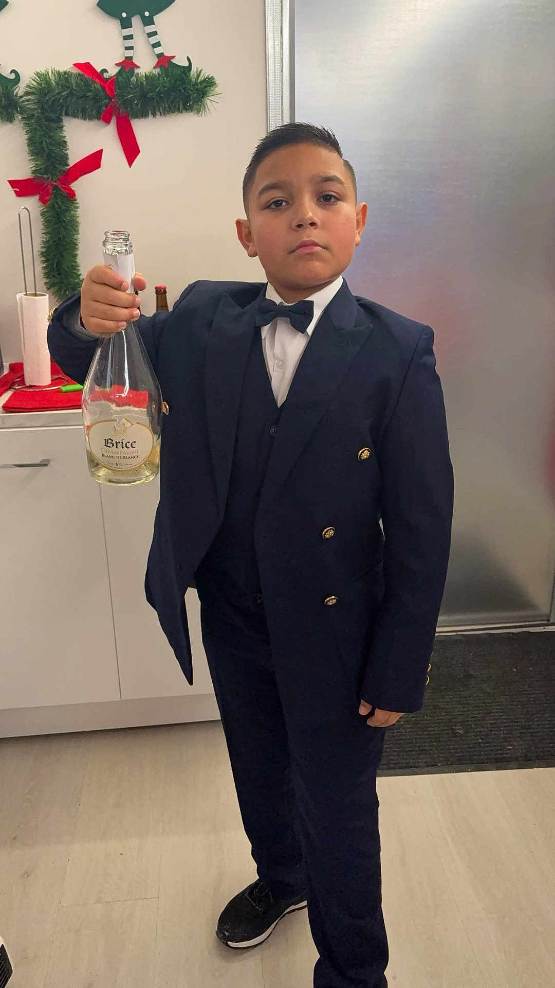 Tony-Junior a rejoint le concours — aidez-le/la à gagner de superbes lots ! boy, child, suit, bow_tie, champagne_bottle, kitchen, holiday_decor, serious_expression, standing, floor, cabinet, door, festive, young, indoors, person, footwear, celebration, portrait, holding