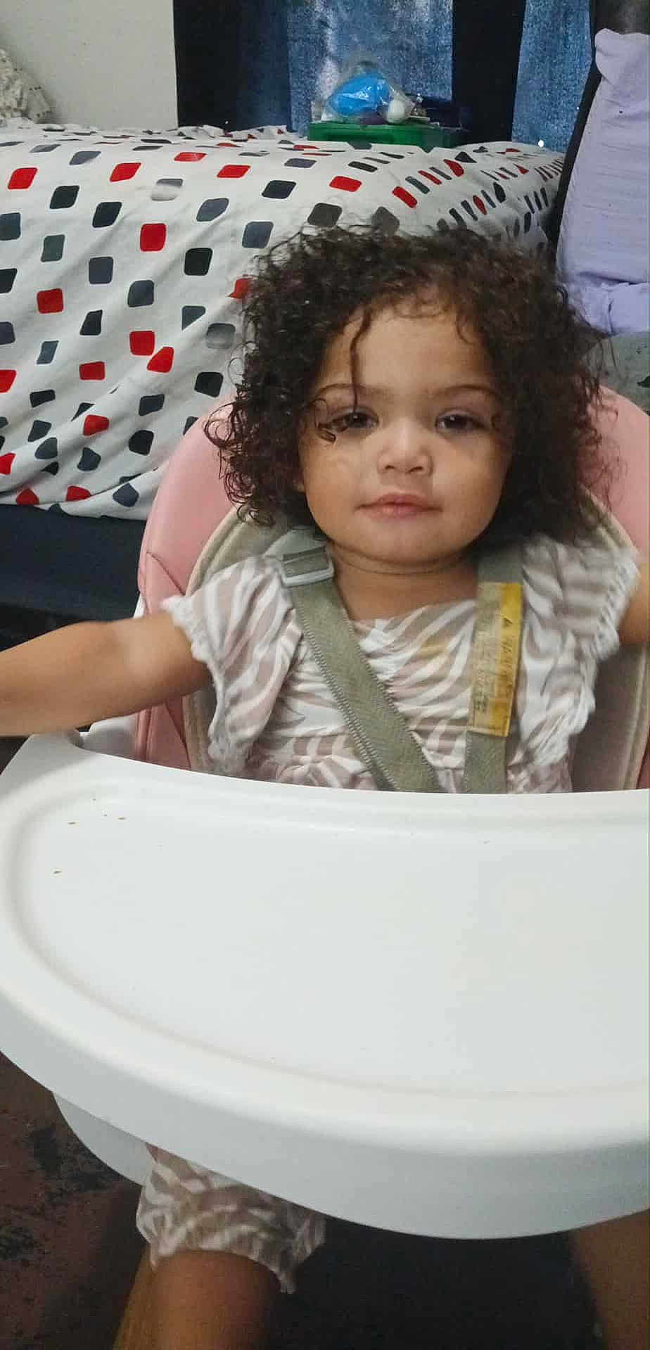 Layla is registered to the contest to win money with this photo: baby, child, clothing, curly_hair, expression, face, furniture, high_chair, home, indoor, infant, patterned_bedspread, person, room, seatbelt, sitting, table, toddler, young_child