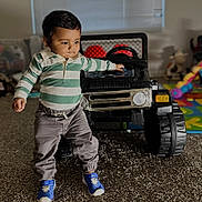 Prayan is registered to the contest to win money with this photo: toddler, child, indoor, toy, jeep, car, playroom, carpet, sneakers, striped_shirt, pants, person, young_child, walking, curious, play, floor, window, plastic_toy, home