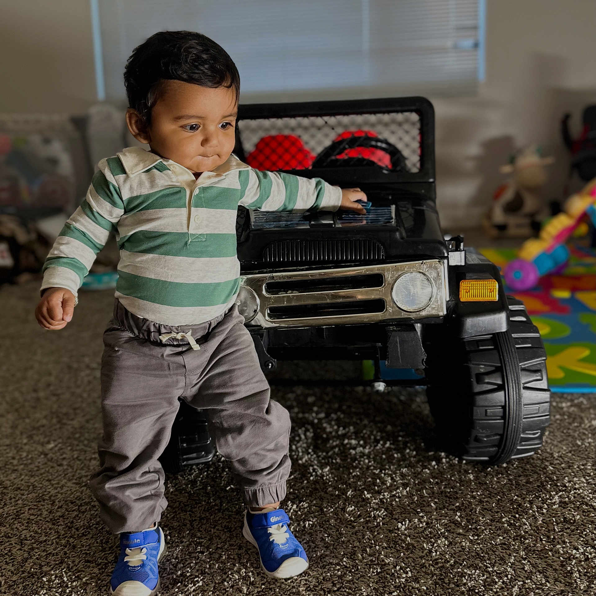Prayan is registered to the contest to win money with this photo: toddler, child, indoor, toy, jeep, car, playroom, carpet, sneakers, striped_shirt, pants, person, young_child, walking, curious, play, floor, window, plastic_toy, home