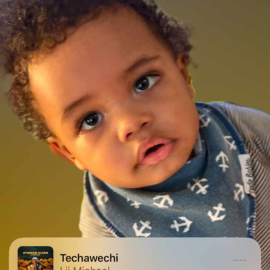 Emmanuel is registered to the contest to win money with this photo: toddler, child, curly_hair, face, blue_bib, striped_shirt, music_player, lock_screen, phone_screen, time_display, date_display, portrait, close_up, indoor, expression, young_child, eyes, head_tilt, casual_clothing, technology