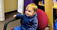 Jackson is registered to the contest to win money with this photo: blue_hoodie, blue_pants, bookshelf, carpeted_floor, casual_clothing, child, curious_expression, furniture, indoor, looking_up, maroon_chair, office, office_chair, person, shelf, sitting, toddler, whiteboard, yellow_shoes, young_child