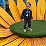 toddler, child, playground, sunflower, yellow_petals, green_center, grass, sweater, shoes, outdoor, fence, play, person, toy, fun, smile, young_child, cute, wood, daylight