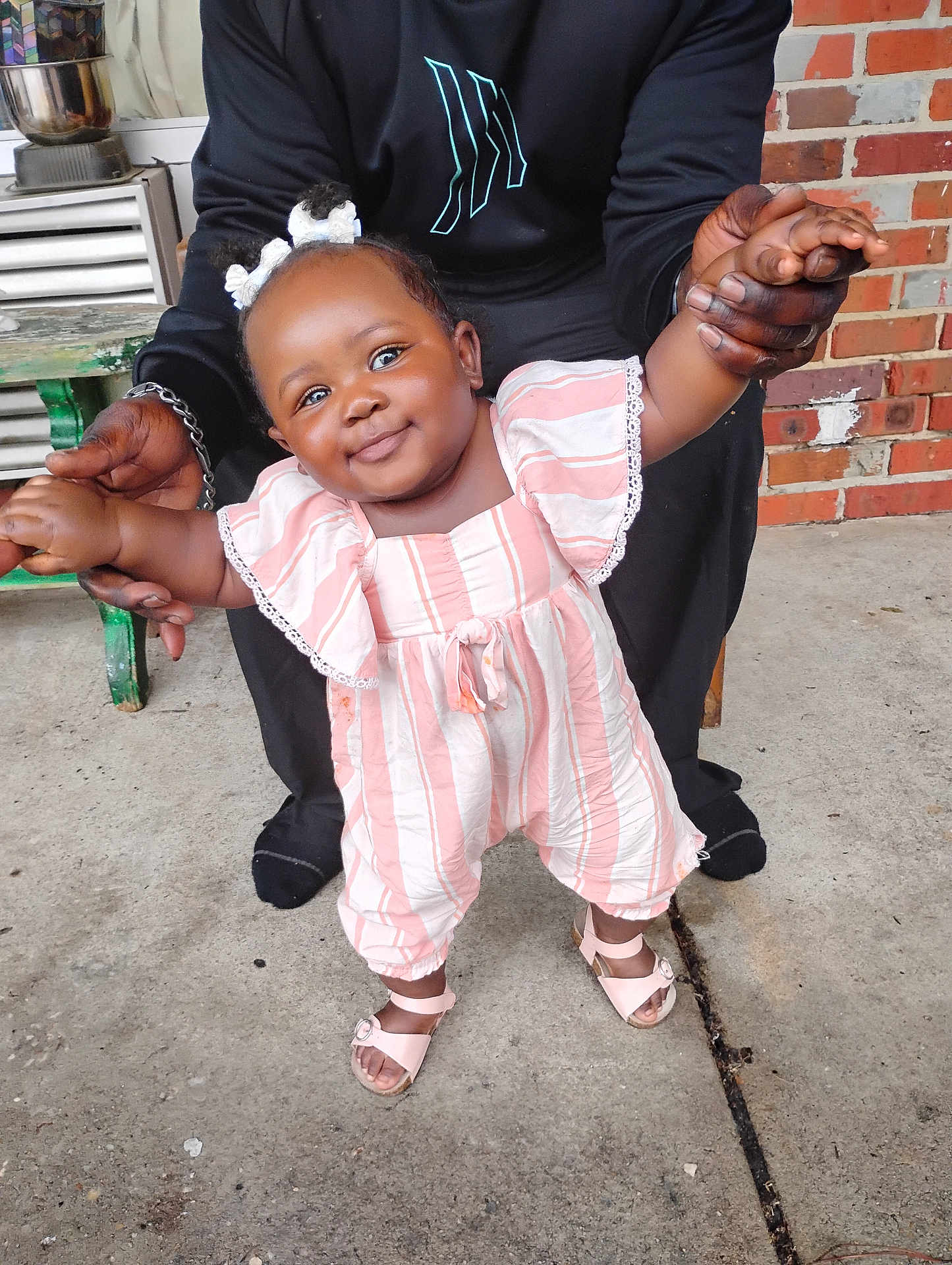 Ariel joined the competition — help win amazing prizes! baby, child, smiling, standing, holding_hands, pink_clothing, striped_clothing, hair_bows, adult_hands, concrete_floor, brick_wall, outdoor, footwear, sandal, cute, person, portrait, happy, support, casual
