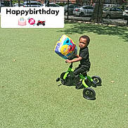 Zion is registered to the contest to win money with this photo: child, tricycle, balloon, outdoor, grass, toy, happybirthday, sesamestreet, green, black_clothing, sunny, daytime, park, play, cute, preschooler, vehicle, smiling, fence, car