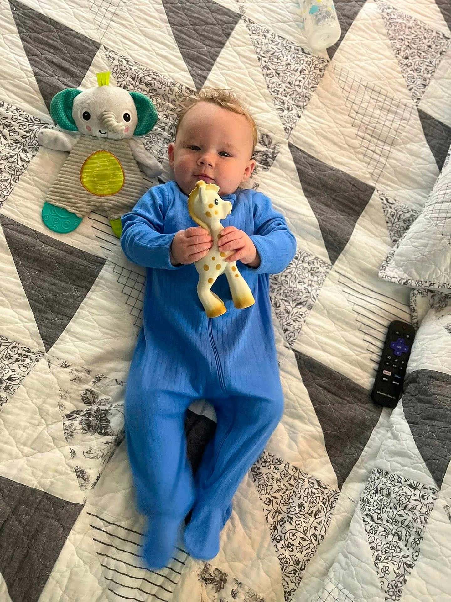 Ashton is registered to the contest to win money with this photo: baby, blue_onesie, giraffe_toy, plush_elephant, baby_bottle, remote_control, quilt, patterned_bedspread, infant, lying_down, toy, child, cute, indoor, bedroom, soft_toys, smiling, person, sleepwear, comfort