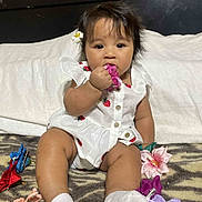 Elel is registered to the contest to win money with this photo: baby, child, white_outfit, strawberry_print, flower_clip, chewing, curious_expression, sitting, blanket, colorful_clips, toy, indoor, cute, infant, person, soft_lighting, head_accessory, happy, small_toys, portrait