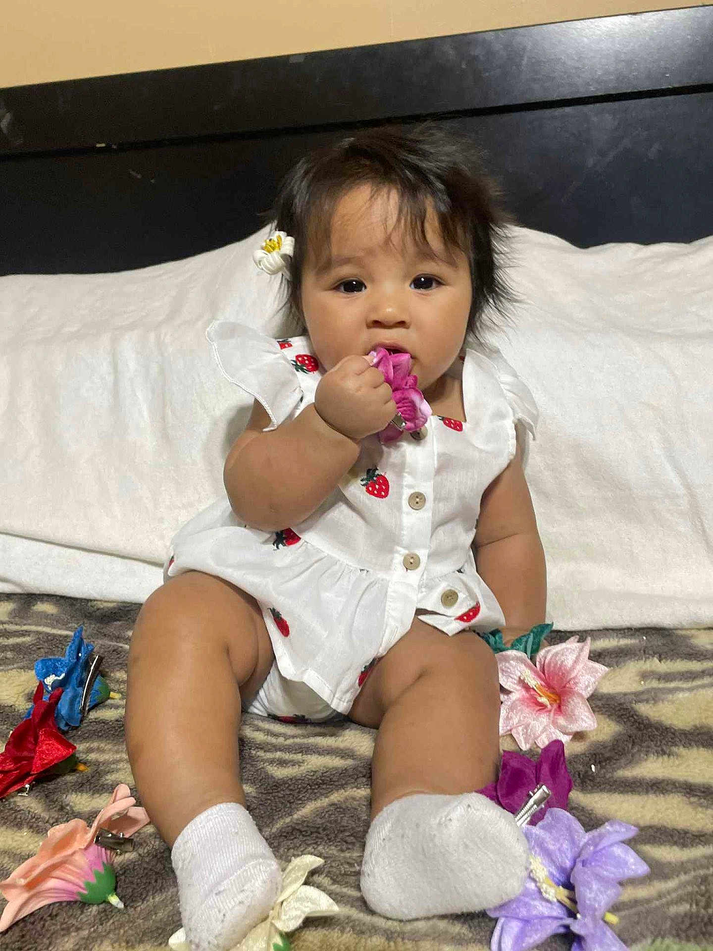 Elel is registered to the contest to win money with this photo: baby, child, white_outfit, strawberry_print, flower_clip, chewing, curious_expression, sitting, blanket, colorful_clips, toy, indoor, cute, infant, person, soft_lighting, head_accessory, happy, small_toys, portrait