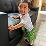 Juan is registered to the contest to win money with this photo: child, toddler, standing, indoor, black_couch, leather, green_shorts, dinosaur_print, white_tshirt, sneakers, floor_tiles, curious_expression, hands_on_couch, short_hair, smiling, home_interior, natural_light, furniture, person, cute