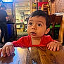 toddler, child, red_shirt, table, wood, indoor, restaurant, curious, face, hands, brown_hair, furniture, sombrero, background, lighting, people, person, casual, expression, candid