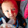 baby, child, pacifier, blue_eyes, stroller, red_background, gray_straps, closeup, infant, curious_expression, holding_object, face, young_child, clothing, seatbelt, indoors, portrait, head, skin, person