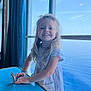 blonde_hair, blue_table, casual, child, compact_mirror, curtain, daylight, girl, happy, indoor, natural_light, ocean_view, person, portrait, reflection, smile, standing, striped_dress, window, young