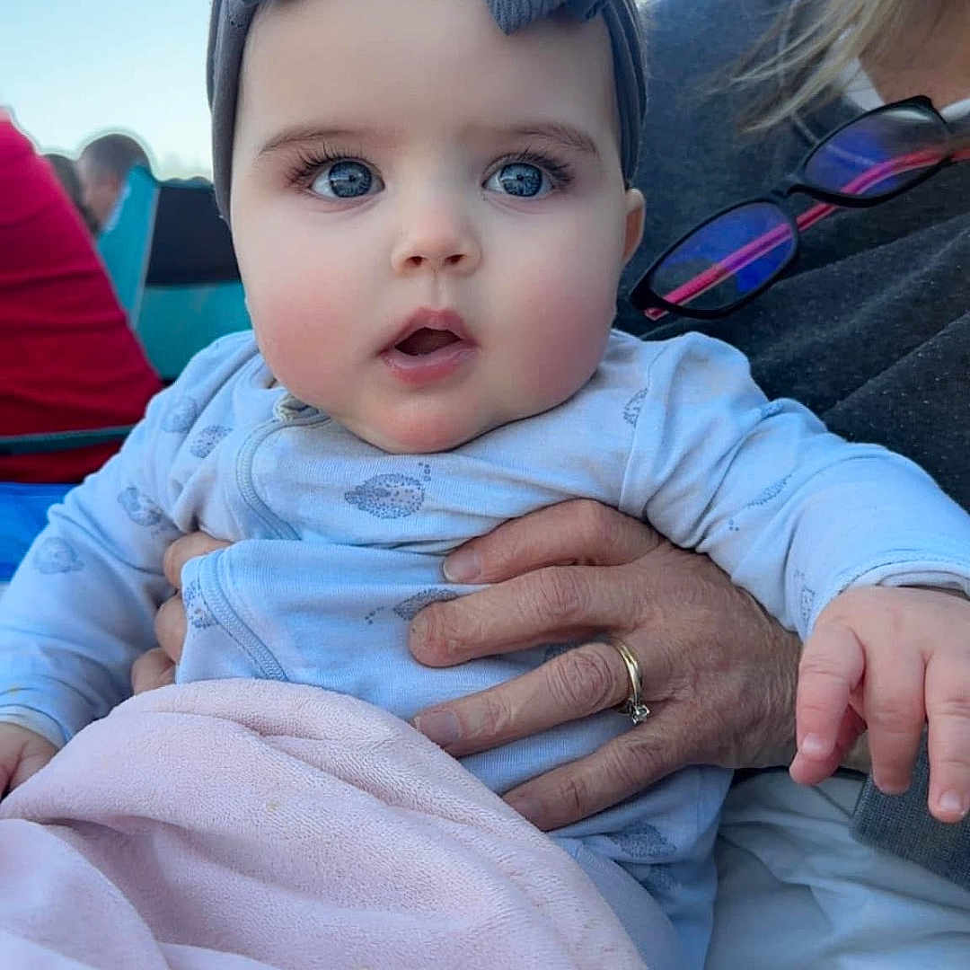 Landynn joined the competition — help win amazing prizes! adult_hand, baby, blanket, blue_eyes, bow, child, clothing, cute, daylight, expression, face, hand, headband, holding, infant, outdoor, person, portrait, sky, warm_clothing