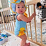 May joined the competition — help win amazing prizes! baby, child, standing, blue_outfit, yellow_duck, baby_hat, wooden_gate, toy, floor_mat, indoor, home, curious, barefoot, cute, infant, play_area, colorful, person, face, expression