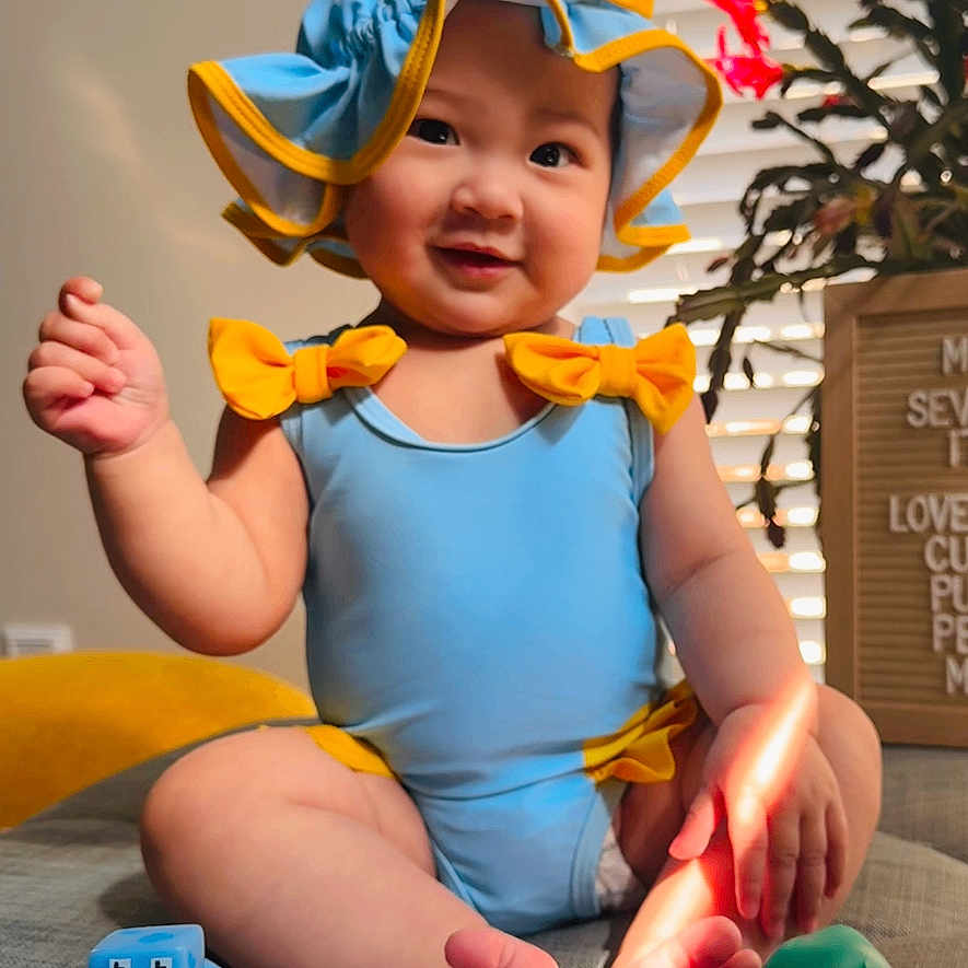 May is registered to the contest to win money with this photo: baby, blue_clothing, yellow_bow, hat, rubber_duck, toy, indoor, smiling, child, sitting, window_blinds, sunlight, colorful, playful, cute, infant, happy, face, foot, home