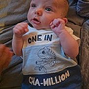 Axel is registered to the contest to win money with this photo: baby, infant, cute, clothing, shirt, text, raised_hands, face, eyes, blanket, cozy, expression, lying_down, skin, newborn, person, portrait, indoors, soft_light, child