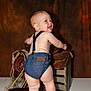 Zion joined the competition — help win amazing prizes! baby, denim_overalls, saddle, wooden_crate, rope, cowboy_gear, barefoot, brown_background, standing, smiling, child, indoor, cute, infant, playful, studio_photo, light_skin, blond_hair, back_view, clothing