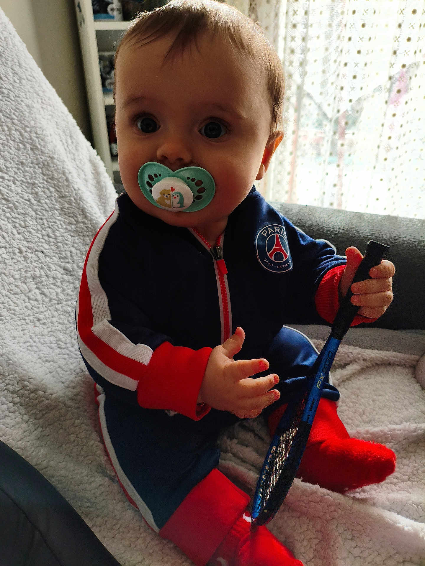 Luka Vucic participe au concours pour gagner de l'argent avec cette photo : baby, pacifier, sportswear, tennis_racket, sitting, blanket, couch, indoor, child, cute, face, clothing, red_socks, blue_jumpsuit, window, curtain, hand, toy, person, portrait