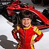 child, boy, smiling, portrait, racing_suit, formula_car, race_car, motorsport, automotive, red, yellow, studio, indoor, standing, hands_on_hips, helmetless, vehicle, sportswear, watermark, capcut_ai