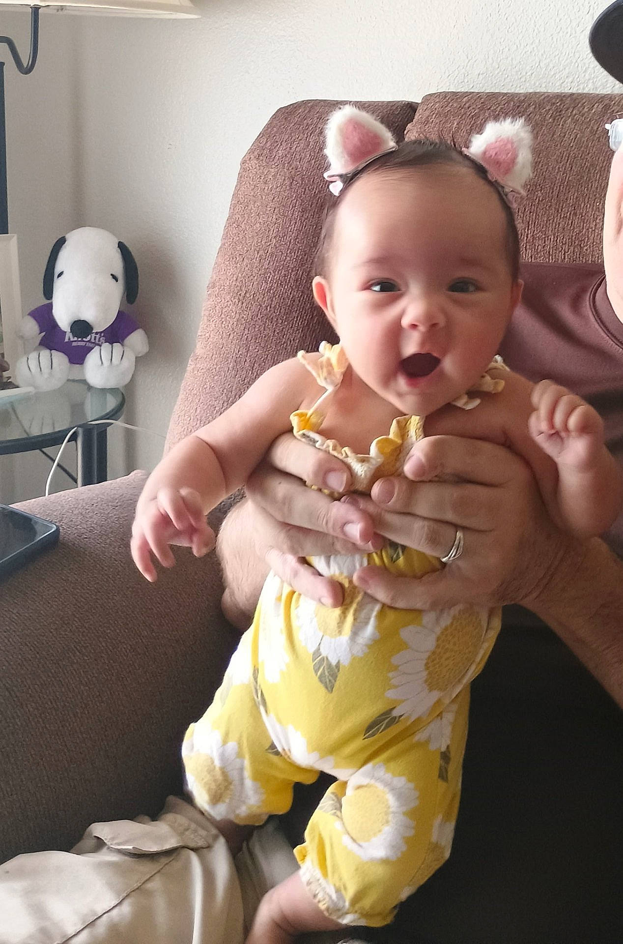 Celine is registered to the contest to win money with this photo: adult_hand, baby, casual, cat_ears, child, couch, cute, flower_pattern, furniture, happy, headband, human, indoor, living_room, person, plush_toy, seated, smiling, snoopy, yellow_outfit