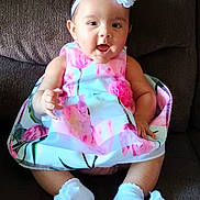 Celine is registered to the contest to win money with this photo: baby, child, girl, dress, floral, headband, flower, socks, white, couch, smiling, happy, seated, indoors, cute, infant, person, apparel, comfort, portrait