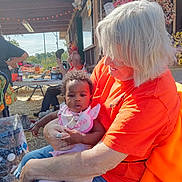 Valerie joined the competition — help win amazing prizes! baby, child, elderly_person, outdoor, daylight, woman, holding, orange_shirt, pink_clothing, wreath, decor, table, chair, person, hat, building, sunlight, nature, people, smile