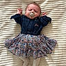 baby, bed, blanket, clothing, cute, denim_jacket, face, floral_skirt, hands_up, head, indoors, infant, lying_down, newborn, pacifier, patterned_fabric, portrait, smiling, soft_texture, tights