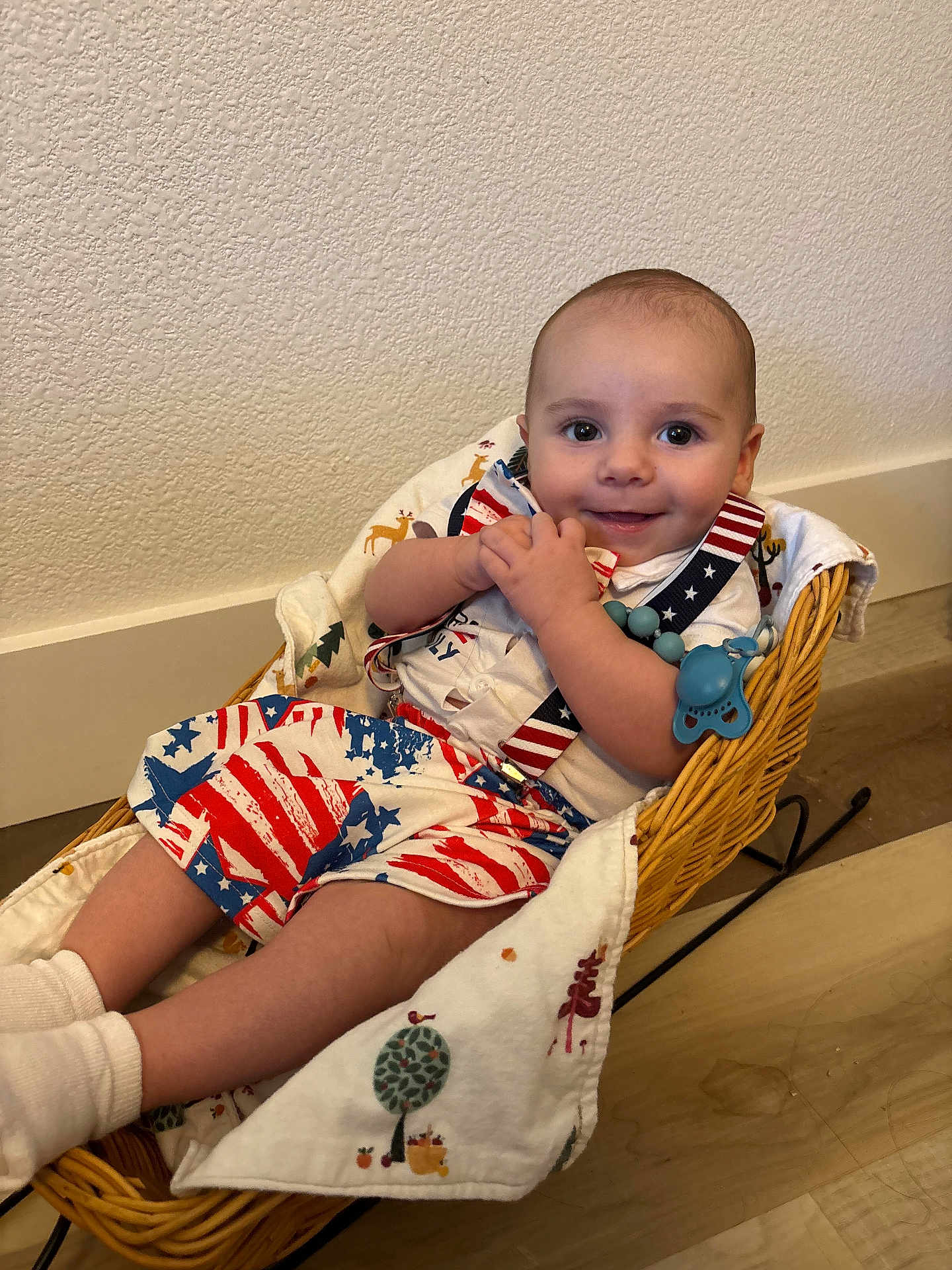 Isaiah is registered to the contest to win money with this photo: baby, smiling, wicker_basket, blanket, suspenders, shorts, indoor, wooden_floor, wall, happy, child, person, clothing, cute, infant, comfort, face, sock, home, relaxing