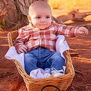 Isaiah joined the competition — help win amazing prizes! baby, casual_clothing, child, curious, cute, daytime, dirt_ground, infant, jeans, nature, outdoor, plaid_shirt, portrait, relaxed, scenic_background, sitting, sunlight, tree_trunk, wicker_basket, young_child