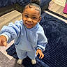 child, toddler, smiling, portrait, indoor, living_room, blue_outfit, hoodie, sweatpants, sneakers, rug, sofa, home, standing, reaching, curly_hair, hair_bun, earrings, floor, photography