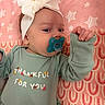 baby, infant, child, portrait, lying_down, pacifier, headband, bow, onesie, clothing, sleeves, hand, raised_arm, blanket, pink, patterned_background, stars, text_on_clothing, indoor, close_up