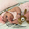 baby, infant, child, portrait, smiling, lying_down, pink_clothing, headband, flower, teddy_bear, stuffed_animal, blanket, bed, pajamas, bracelet, indoors, close_up, happy, playful, cozy
