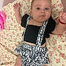 baby, infant, child, portrait, lying_down, wide_eyed, facial_expression, smiling, arms_raised, floral_blanket, black_outfit, patterned_clothing, suspenders, skirt, pacifier, bed, indoor, close_up, adorable, photography