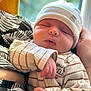 newborn, baby, sleeping, hat, striped_clothing, hand, arm, person, closeup, window, blurred_background, indoor, cozy, soft_lighting, peaceful, infant, cute, resting, portrait, toddler