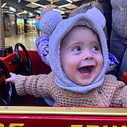 Tiago a rejoint le concours — aidez-le/la à gagner de superbes lots ! adult, amusement_ride, baby, bear_hood, candid, child, hand_on_wheel, hat, indoor, joyful, knit_sweater, laughing, person, red_car, shopping_mall, smiling, sparkly_lights, steering_wheel, toddler, toy