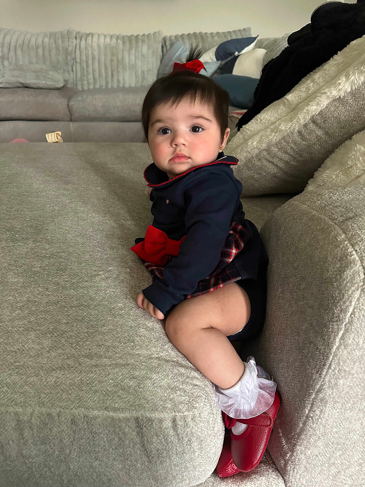 Isabella joined the competition — help win amazing prizes! baby, child, girl, couch, sofa, red_bow, red_shoes, white_socks, plaid_skirt, navy_top, living_room, cushion, soft_furniture, indoors, portrait, sitting, curious, cute, young_child, home